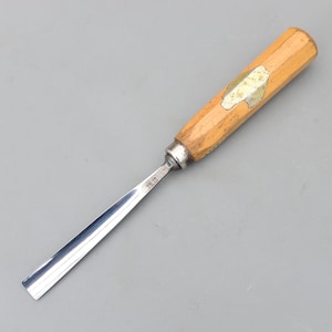 以下が含まれることがあります： 光沢のある鋼の刃と明るい色の木製ハンドルを備えた木彫りのノミ。 ハンドルには金属製のフェルールと小さな装飾要素があります。 このツールは、木工や彫刻用に設計されています。