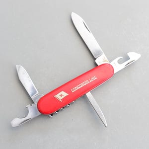 May include: A red pocket knife with a white flag featuring a blue "H" and the words "CONCORDIA LINE" on the handle. The knife includes a bottle opener, a small blade, a larger blade, and a saw.