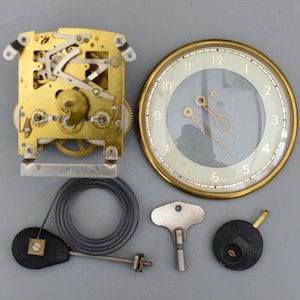 May include: Clock parts including a gold-toned clock face with hands, a brass movement, a key, a pendulum, and a coiled spring. The clock face has a light green background with black numerals.