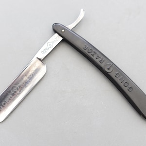 Puede incluir: Una navaja de afeitar abierta con mango negro y hoja plateada. La hoja está grabada con las palabras "GONG RAZOR" y "HOLLOW GROUND". El mango es curvo y tiene las palabras "GONG RAZOR" impresas.