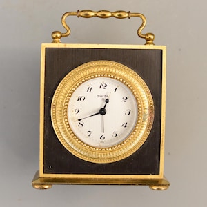 Può includere: Un orologio quadrato nero e dorato con quadrante bianco. L'orologio ha una maniglia dorata e una cornice dorata attorno al quadrante. Il quadrante ha lancette nere e la parola "SWIZA" stampata sopra.