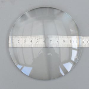 以下が含まれることがあります： 透明な円形の拡大レンズに定規が置かれています。定規はセンチメートルで測定し、レンズの直径を示しています。レンズは透明で、周囲の環境を反射しています。画像はニュートラルグレーの表面で撮影されています。