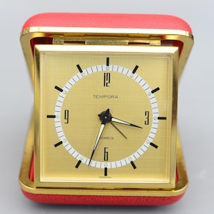 Peut inclure: Un réveil carré de couleur or avec un cadran blanc et des aiguilles noires. L'horloge est dans un étui en cuir rouge et porte les mots "Tempora" et "Jewels" sur le cadran.