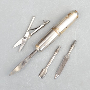 Puede incluir: Un juego de manicura vintage con un estuche de metal plateado y varias herramientas. El juego incluye tijeras pequeñas, un empujador de cutículas, una lima de uñas y una cuchilla. Las herramientas están dispuestas sobre una superficie gris.