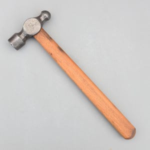 May include: A ball-peen hammer with a wooden handle. The hammer head is made of metal and has a rounded striking face on one side and a ball-shaped peen on the other. The handle is a light brown color.