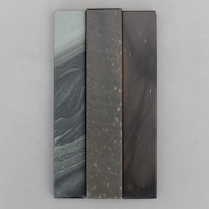 May include: Three rectangular blocks arranged vertically side by side. The left block features a grey and black swirling pattern. The center block is a solid matte grey with light speckles. The right block is a smooth, dark brown with subtle shading.