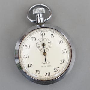 Può includere: Un cronometro Smiths vintage con cassa color argento e quadrante bianco. Il quadrante presenta numeri neri, un quadrante più piccolo per i minuti e la scritta "SMITHS 1/5TH SECS". Il cronometro ha una corona e un anello.