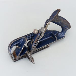 Op de afbeelding: Een blauwe metalen handvlak met een houten handvat. De vlak heeft een metalen mes en een metalen instelknop.