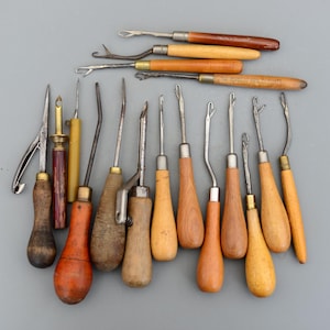 Puede incluir: Una colección de 17 herramientas vintage con mango de madera y puntas de metal. Las herramientas tienen diferentes formas y tamaños, incluyendo ganchos, agujas y punzones. Algunas de las herramientas tienen detalles en latón. Las herramientas están dispuestas sobre una superficie gris.