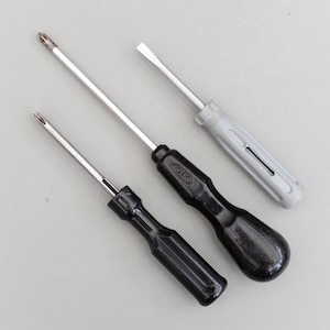 Może przedstawiać: Trzy śrubokręty ze srebrnymi wałkami i uchwytami w różnych kolorach. Dwa mają czarne uchwyty, a jeden szary. Końcówki śrubokrętów to mieszanka typów płaskich i krzyżakowych. Narzędzia są ułożone po przekątnej.