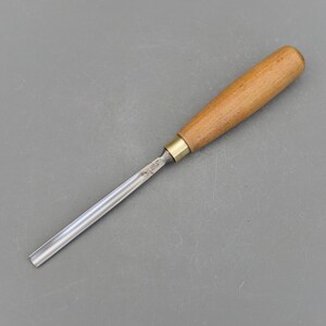 以下が含まれることがあります： 木製のハンドルと金属製の刃を持つ木彫りツール。刃は薄くて平らで、ハンドルは茶色です。
