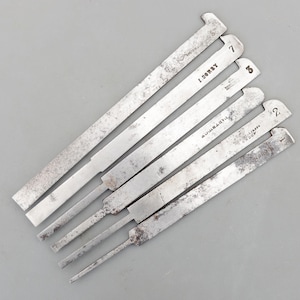 May include: A collection of seven antique metal tools, each with a rectangular body and a notched end. The tools are arranged diagonally, displaying varying degrees of wear and tear. Numbers 1 through 8 are visible, along with the names of the manufacturers.
