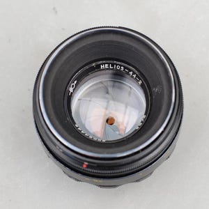 May include: A black Helios-44-2 camera lens with a silver interior. The lens has a series of markings and numbers around the edge, including the text "Helios-44-2". The lens has a red dot and a series of aperture blades visible inside.