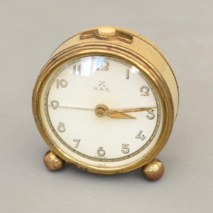 Op de afbeelding: Een vintage, ronde wekker met een crèmekleurige wijzerplaat en een goudkleurige metalen behuizing. De wijzerplaat heeft zwarte cijfers en wijzers, met de tekst "H.A.C." en "FOREIGN". De klok heeft twee kleine, ronde voetjes.