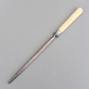 以下が含まれることがあります： クリーム色のハンドルと銀色の金属製のボルスターが付いた、ヴィンテージのスチール製ナイフシャープナー。シャープナーは長く先細りのスチールロッドで、ハンドルは円筒形です。「CAST STEEL」の文字がスチールロッドにエッチングされています。