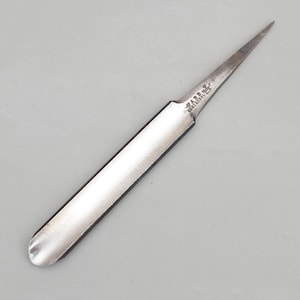 May include: A metal gouge tool with a curved, concave blade and a pointed tip. The metal surface shows signs of wear and age. The text WARD CAST STEEL is stamped clearly on the metal near the transition from the handle area to the pointed tip.