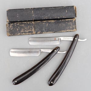 Może przedstawiać: Dwa antyczne brzytwy z czarnymi rękojeściami i srebrnymi ostrzami. Brzytwy znajdują się w oryginalnych czarnych skórzanych etui. Ostrza są oznaczone "MANUFACTURED FOR" i "SHEFFIELD, ENGLAND".
