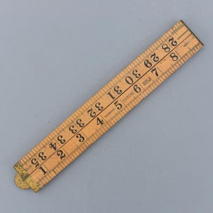 Pode incluir: Uma régua dobrável de madeira vintage, medindo até 89 cm. A régua é feita de madeira clara com marcações e números pretos. A régua é marcada "RABONE" e "MADE IN ENGLAND". A régua está dobrada e tem uma dobradiça de latão.