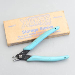 Puede incluir: Un cortador de alambre azul de la marca Xuron con un mango negro y el texto "WIRE SAFETY" en el mango. Los cortadores están en una caja marrón con el texto "XURON CORP. Storage Sleeve" en la caja.