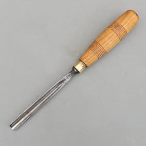 Könnte beinhalten: Ein Holzbeitel mit einem hellbraunen Holzgriff und einer silbernen Metallklinge. Der Griff hat horizontale Rillen. Das Werkzeug dient zum Formen und Schnitzen von Holz. Der Griff ist etwa 10 cm lang.