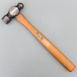 Puede incluir: Un martillo de bola con mango de madera y cabeza de metal oscuro. La cabeza tiene una cara de golpe redonda y una pena en forma de bola. El mango es de color marrón claro y una etiqueta está adherida al mango. El martillo es un Stanley BS 876 WLS.