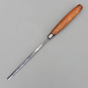 Puede incluir: Un formón para trabajar la madera con mango de madera y hoja de metal. El mango es de color marrón claro y la hoja es plateada. El formón está diseñado para dar forma y tallar madera. La herramienta está en ángulo diagonal.