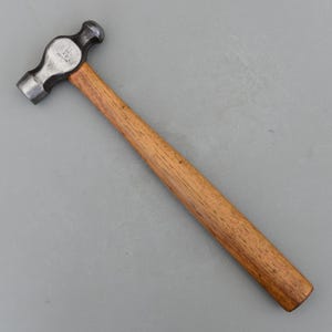 May include: A ball-peen hammer with a wooden handle and a steel head. The hammer head is dark gray and the handle is a light brown color. The hammer is angled from the top left to the bottom right.