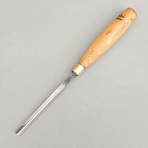 May include: A wood carving gouge featuring a long, semi-circular metal blade with a polished finish and a light brown wooden handle. A brass ferrule connects the metal shaft to the handle, which displays a visible wood grain and dark markings.