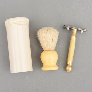 Op de afbeelding: Een vintage scheerset met een crèmekleurige cilindrische container, een scheerkwast met een crèmekleurige handgreep en een veiligheidsscheermes met een crèmekleurige handgreep en zilveren mes. De set staat op een grijze ondergrond.