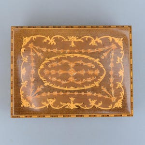 Könnte beinhalten: Eine Holz-Spieluhr mit einem aufwendigen Intarsienmuster aus Blumen und Spiralen. Die Box ist aus hellem und dunklem Holz gefertigt und hat eine rechteckige Form.