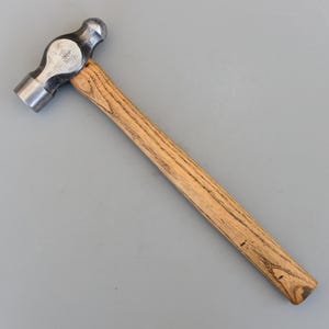 Può includere: Un martello a palla con manico in legno e testa in acciaio. La testa del martello ha una penna arrotondata su un lato e una faccia piatta sull'altro. Il manico è in legno marrone chiaro.