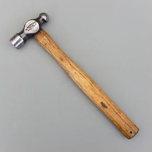 Könnte beinhalten: Ein Kugelkopfhammer mit Holzgriff. Der Hammerkopf besteht aus Metall und hat eine abgerundete Schlagfläche. Der Griff ist aus Holz und hat eine natürliche Oberfläche. Der Hammer ist diagonal auf einer grauen Oberfläche angeordnet.