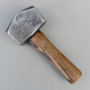May include: A vintage steel hammer with a wooden handle. The hammerhead is rectangular with beveled edges and stamped with the text "SOLICAS STEEL". The wooden handle has a natural wood grain pattern.