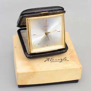Puede incluir: Un reloj despertador de viaje Kienzle vintage en un estuche de cuero negro. El reloj tiene un marco dorado, una esfera plateada y agujas doradas. El reloj está sobre una caja beige con el logotipo de Kienzle.