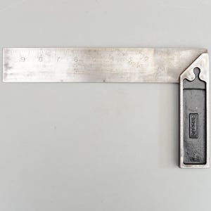 Pode incluir: Um esquadro de metal vintage com uma régua marcada em centímetros. O esquadro tem uma alça retangular escura com a marca "RABONE" visível. A régua estende-se até 23 cm e mostra sinais de desgaste.