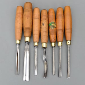 May include: A collection of eight wood carving chisels with wooden handles and metal blades. The chisels have brass ferrules and vary in blade shape, including straight, curved, and angled designs. The handles are a light brown color.