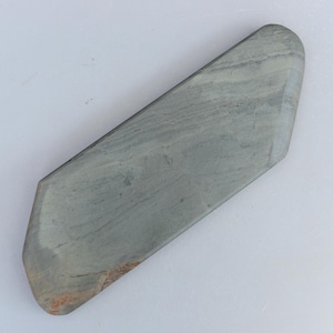 Pode incluir: Uma pedra de amolar cinza com formato retangular e bordas anguladas. A pedra tem uma superfície lisa com padrões de grãos visíveis e uma ligeira variação de cor. A pedra tem aproximadamente 20 cm de comprimento.