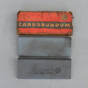 Może przedstawiać: Dwa kamienie szlifierskie Carborundum, jeden oznaczony "No. 183 FINE", a drugi nieoznaczony, w oryginalnym czerwonym pudełku.