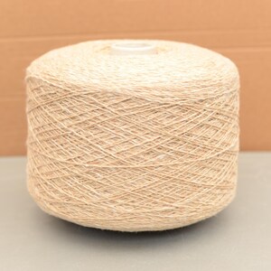 May include: A large, cylindrical spool of beige yarn. The yarn is tightly wound, creating a textured surface. The spool is centered against a neutral background, highlighting the natural colour and texture of the yarn. Suitable for knitting and crochet projects.