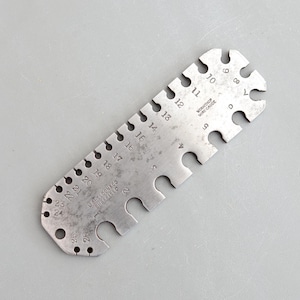 Puede incluir: Un calibrador de alambre de metal vintage con una serie de orificios y recortes numerados. El calibrador tiene el texto "WORKSHOP WIRE GAUGE" y "U SHELDON & CO SHEFFIELD" estampado. Los números van del 1 al 26.