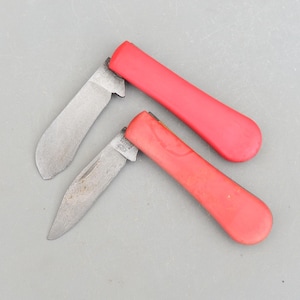 Puede incluir: Dos navajas de bolsillo con mangos rojos y hojas de metal. Las hojas están cerradas.