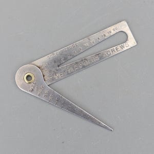 May include: A vintage metal screw gauge by Nettlefolds Screws. The silver-toned gauge features measurements for screw sizes, including "SCREW GAUGE" and "SIZE OF HOLE SG". This tool is designed to measure screw sizes and is used for woodworking and metalworking.