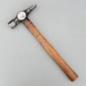 May include: A metal hammer with a wooden handle. The hammerhead has a flat face and a curved claw. The handle is made of light brown wood. The hammer is designed for construction and repair work.