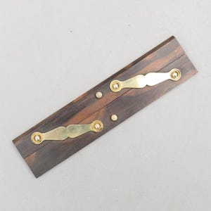 Può includere: Un oggetto rettangolare in legno con finitura marrone scuro, caratterizzato da due elementi decorativi color ottone e piccoli rivetti in ottone. Gli elementi in ottone hanno un design curvo e ornato. L'oggetto è posto su uno sfondo grigio neutro.