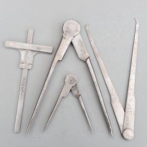 May include: A collection of antique metal measuring instruments, including calipers, dividers, and a T-square. The tools are made of a silver-coloured metal and are laid out on a grey surface. The tools show signs of use and wear.