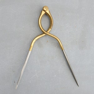 Puede incluir: Un compás de latón con acabado dorado y dos patas puntiagudas. El compás está abierto y listo para usar.