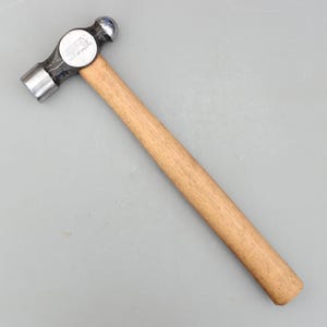 May include: A ball-peen hammer with a wooden handle and a metal head. The hammer head has a flat face on one side and a rounded, ball-shaped face on the other. The handle is made of light brown wood.
