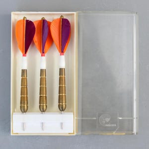 Peut inclure: Trois fléchettes avec des plumes orange et violettes, des tiges blanches et des corps en laiton sont présentées dans un étui en plastique transparent. Les fléchettes sont disposées dans un support blanc à l'intérieur de l'étui. L'étui est ouvert.
