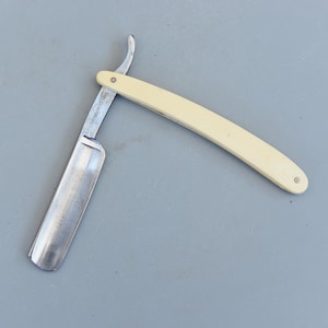 Puede incluir: Una navaja de afeitar recta vintage con un mango color crema y una hoja plateada. La hoja está abierta, revelando el filo. La navaja está sobre una superficie gris.