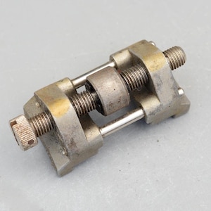 May include: A vintage metal tool with a silver finish. The tool has a rectangular frame with threaded screws and a central cylindrical component. The tool appears to be used for sharpening or holding objects. The tool is on a grey background.